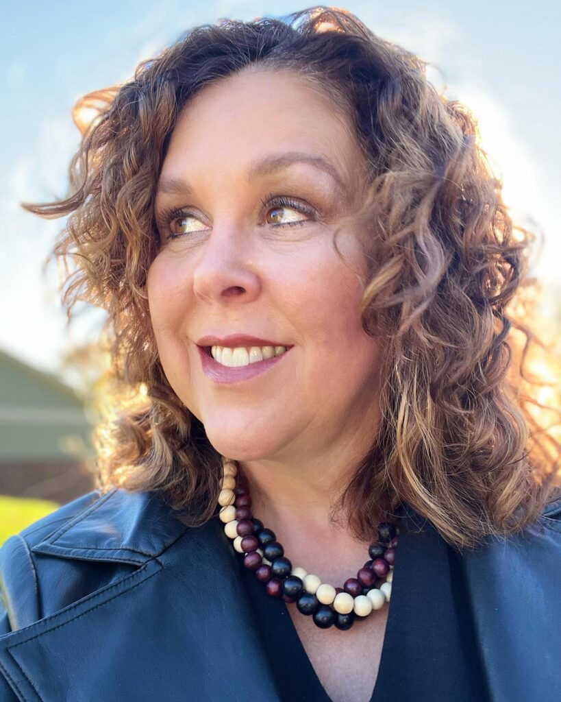 A smiling woman with medium-length curly brunette hair featuring warm caramel highlights and plenty of natural volume.