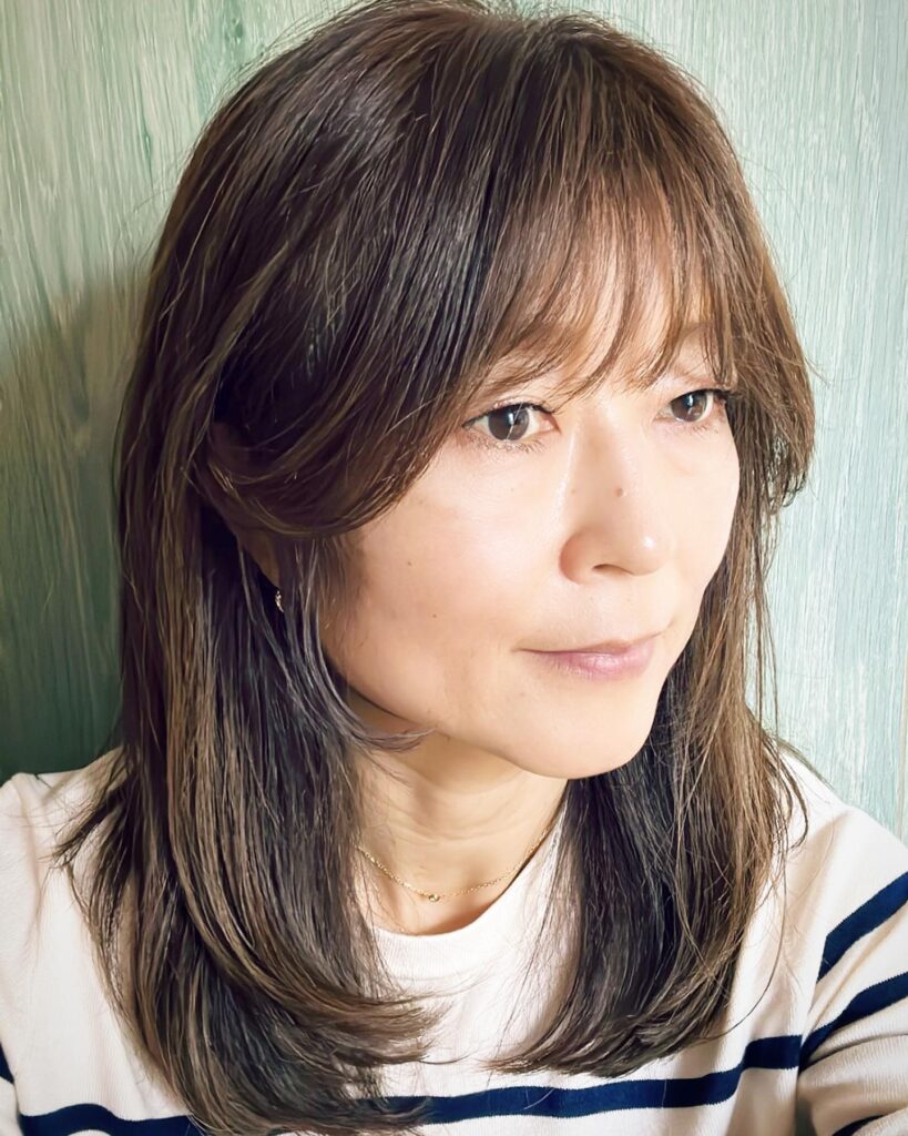 A woman with shoulder-length mocha brown hair styled with soft layers and wispy see-through bangs.