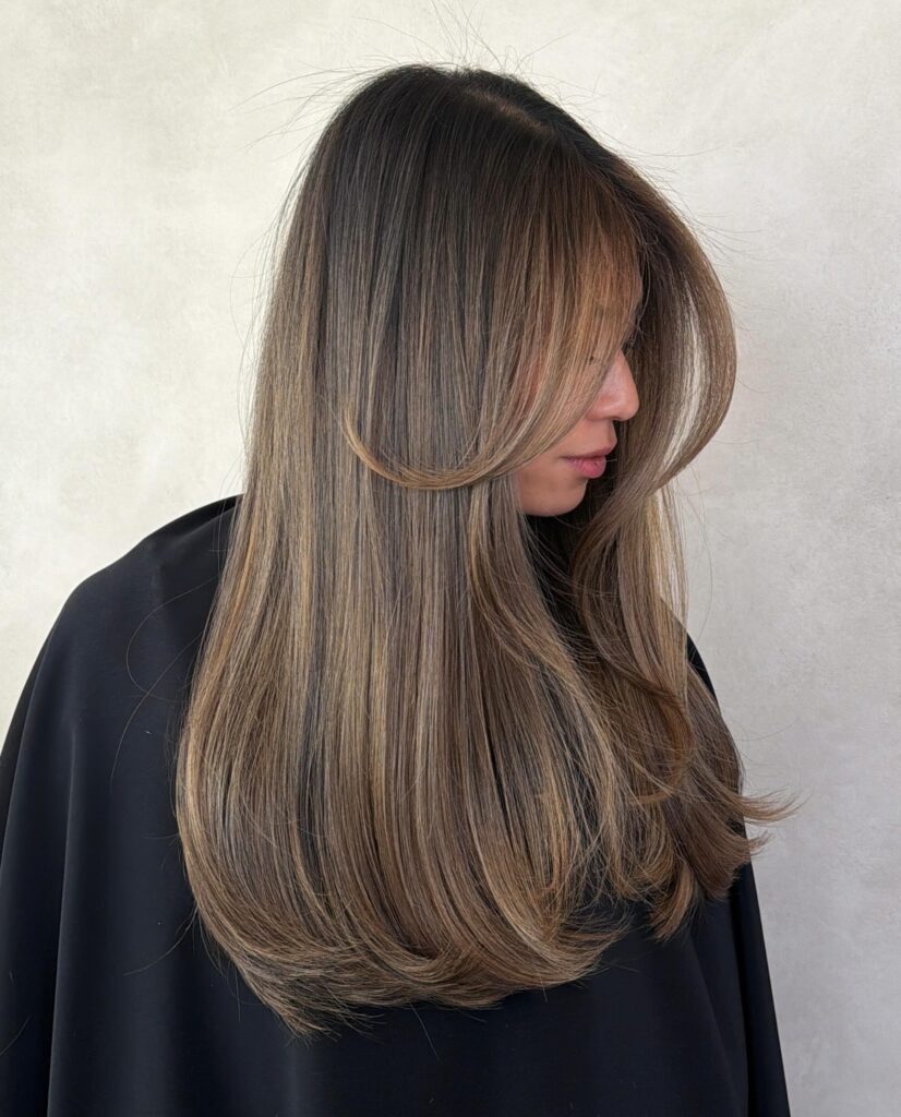 A woman with long straight dark hair featuring fine mushroom bronde highlights and long sweeping curtain bangs shown in profile.
