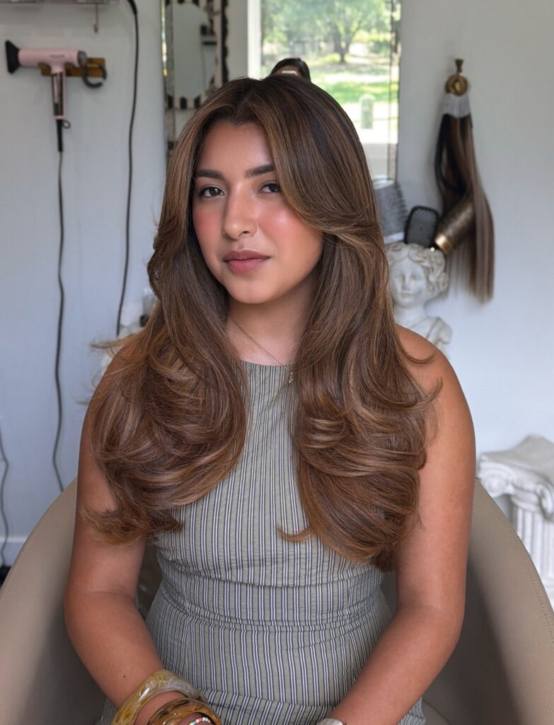 A young woman with long thick bronde hair styled in a bouncy layered blowout looks softly at the camera.