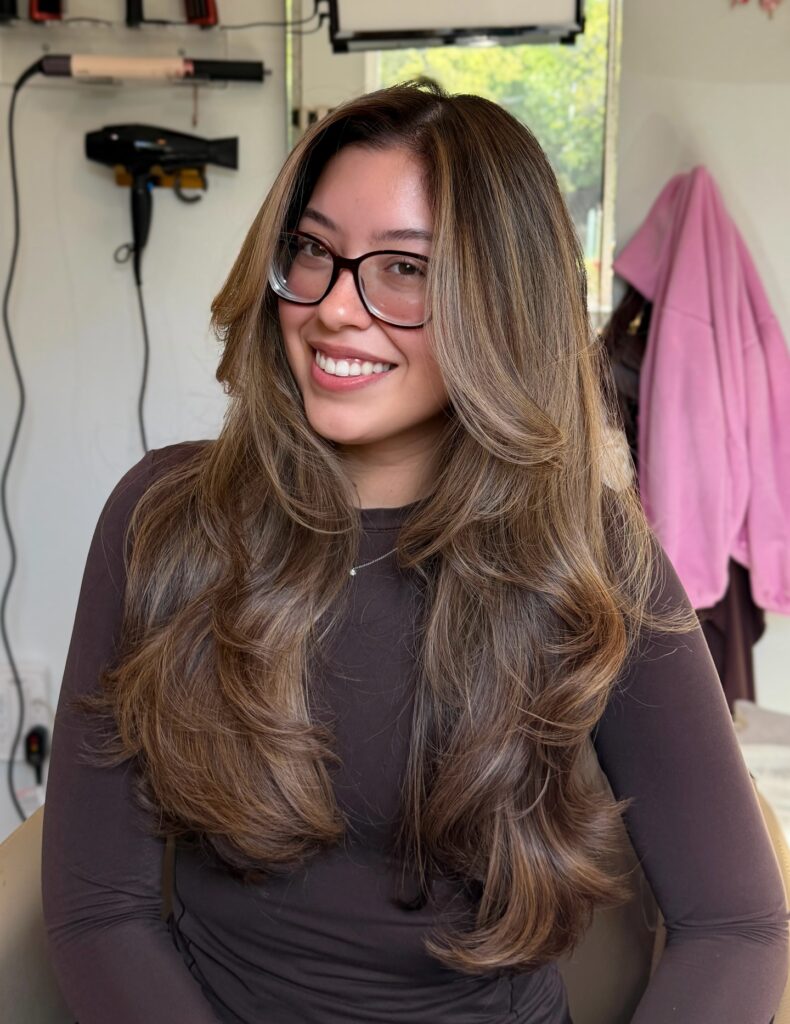 A smiling woman with long layered brown hair with golden highlights and bouncy waves looks forward.