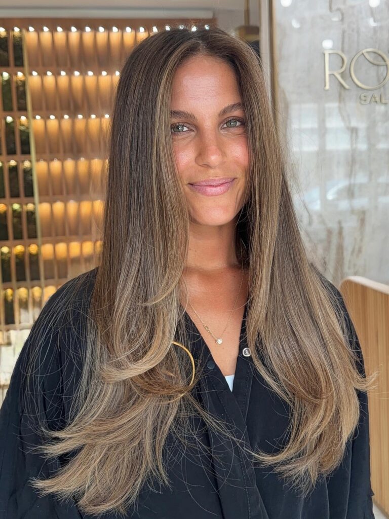 A smiling woman with long straight light brown hair featuring fine beige blonde highlights and soft face framing layers.