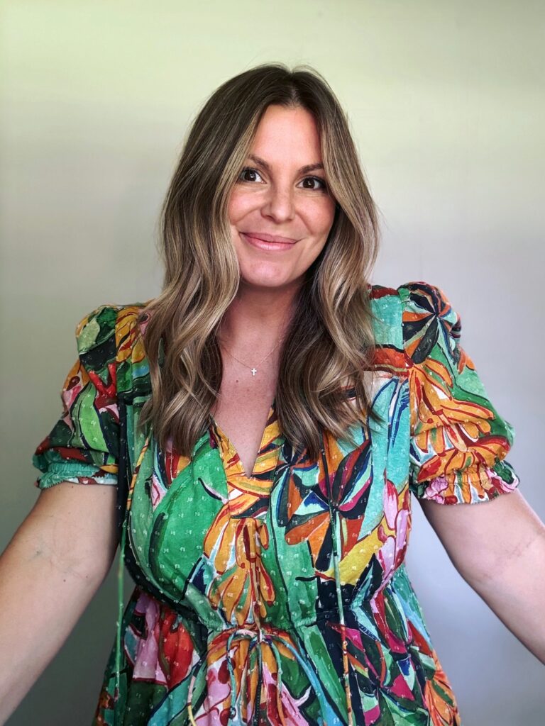 A smiling woman in a colorful floral dress wearing her medium length brown hair in loose waves with golden blonde highlights.