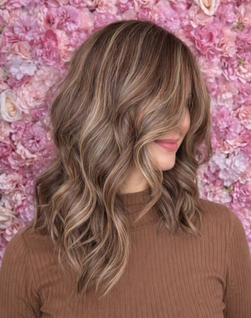 A woman with shoulder length wavy hair displaying soft sandy blonde highlights against a backdrop of pink flowers.