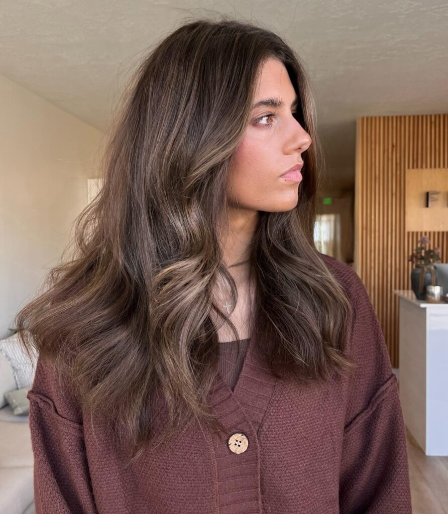 A young woman with long wavy ash brown hair styled with soft cool blonde highlights and face-framing layers.