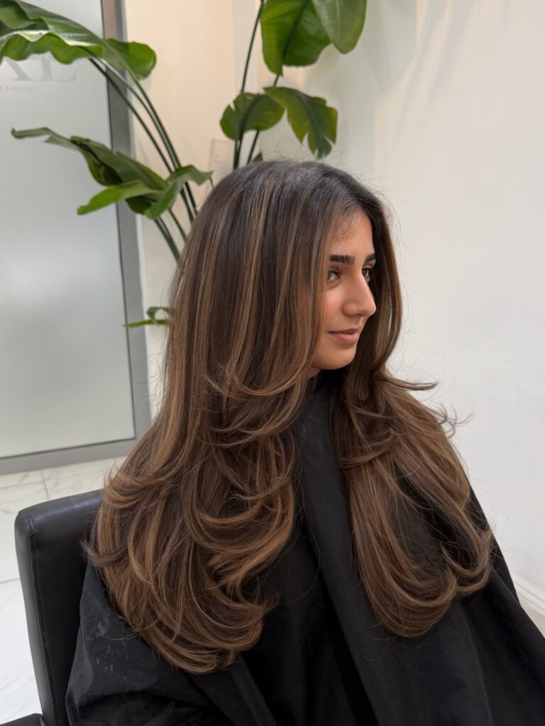 A young woman sitting in a salon chair showcases her long chestnut brown hair styled in bouncy feathery layers with subtle bronde highlights.