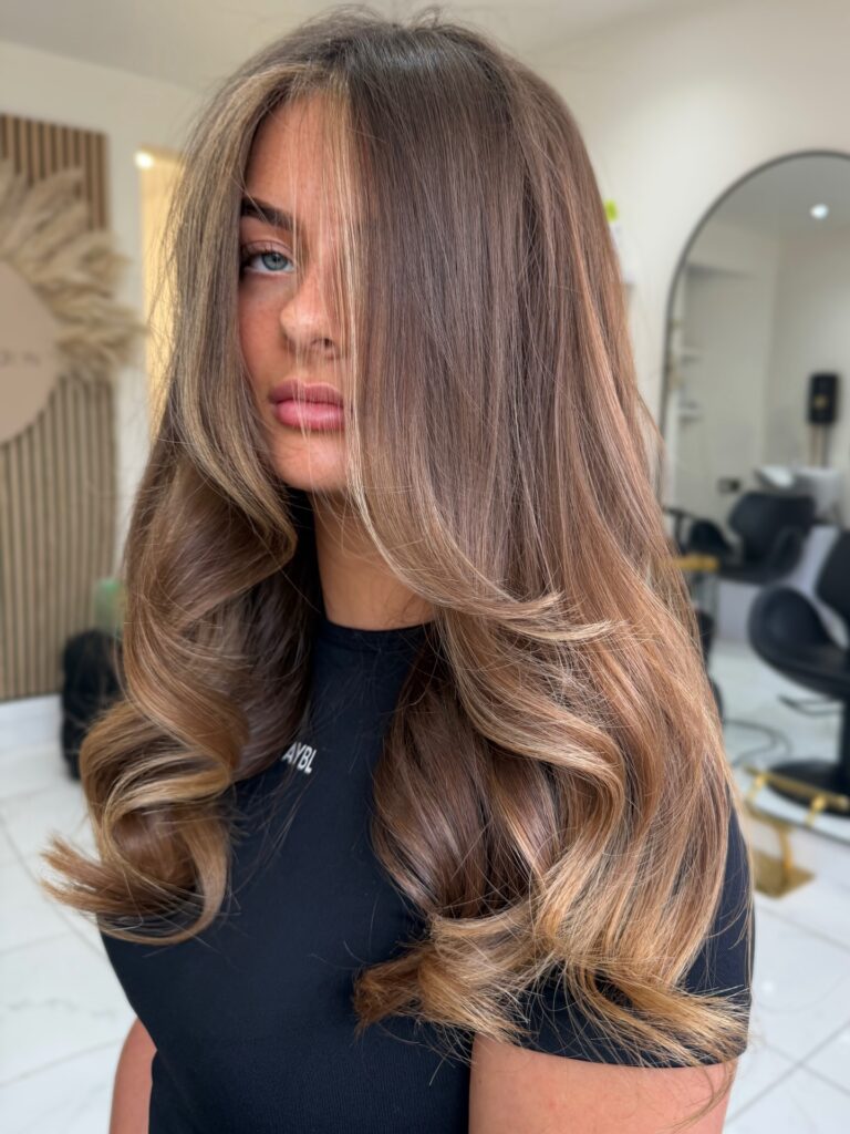 A young woman with smooth, voluminous long ash brown hair featuring subtle blended lighter highlights and soft, bouncy waves focused on the ends.