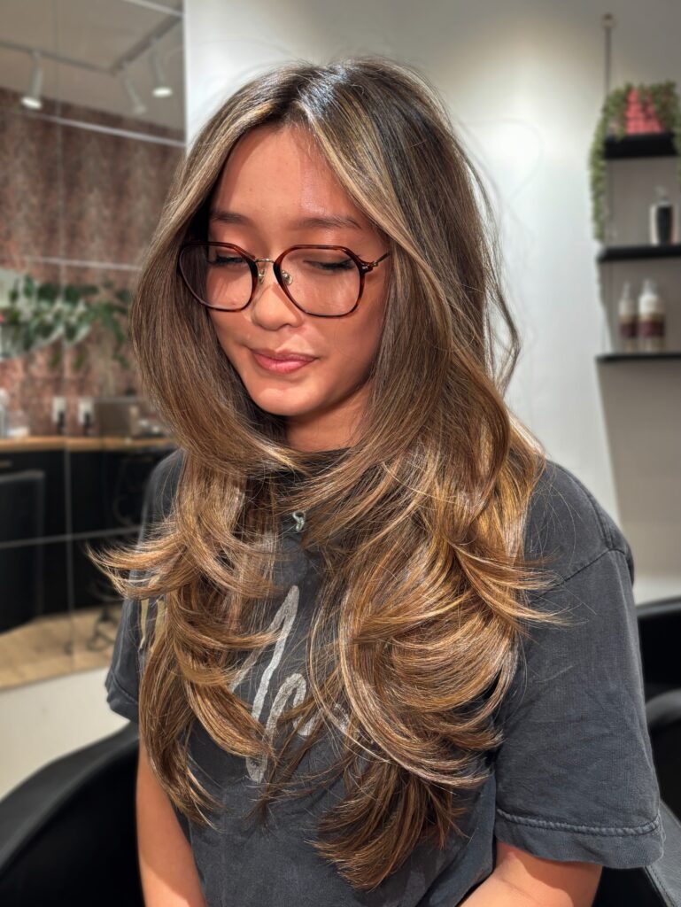 A smiling woman with long layered brunette hair featuring bright, dramatic golden blonde face framing balayage and voluminous curled ends.