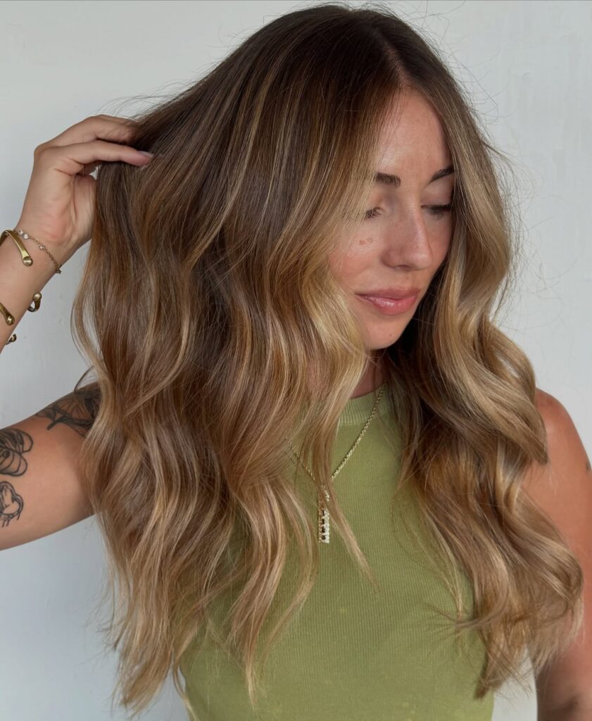 A woman with long wavy honey blonde hair featuring bright face-framing highlights and a natural root blend.