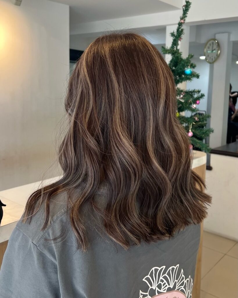 A young woman with shoulder-length wavy dark brown hair featuring blended caramel highlights and subtle layers.