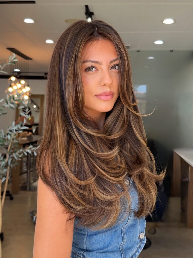 A beautiful woman with long brown hair styled in bouncy layers with warm golden highlights and face-framing bang pieces.