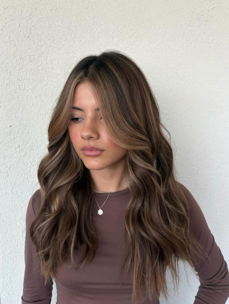 A young woman with long wavy mocha brown hair featuring subtle blonde highlights and bright face-framing curtain layers.