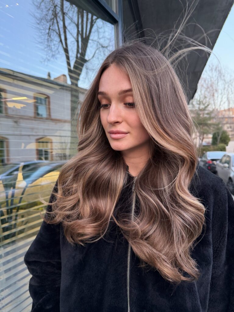 A young woman with long wavy ash bronde hair featuring cool beige highlights stands outside in natural light.