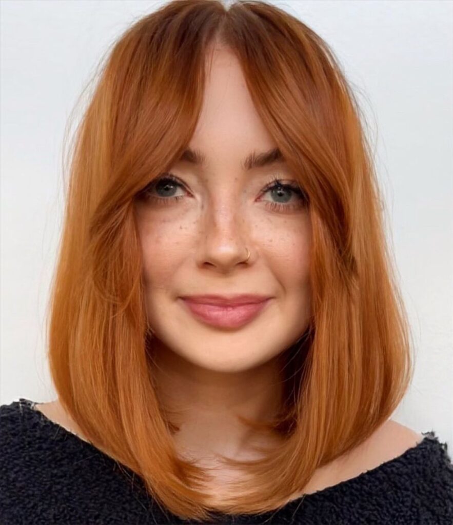 A woman with a bright copper collarbone length lob haircut featuring long curtain bangs and a center part.