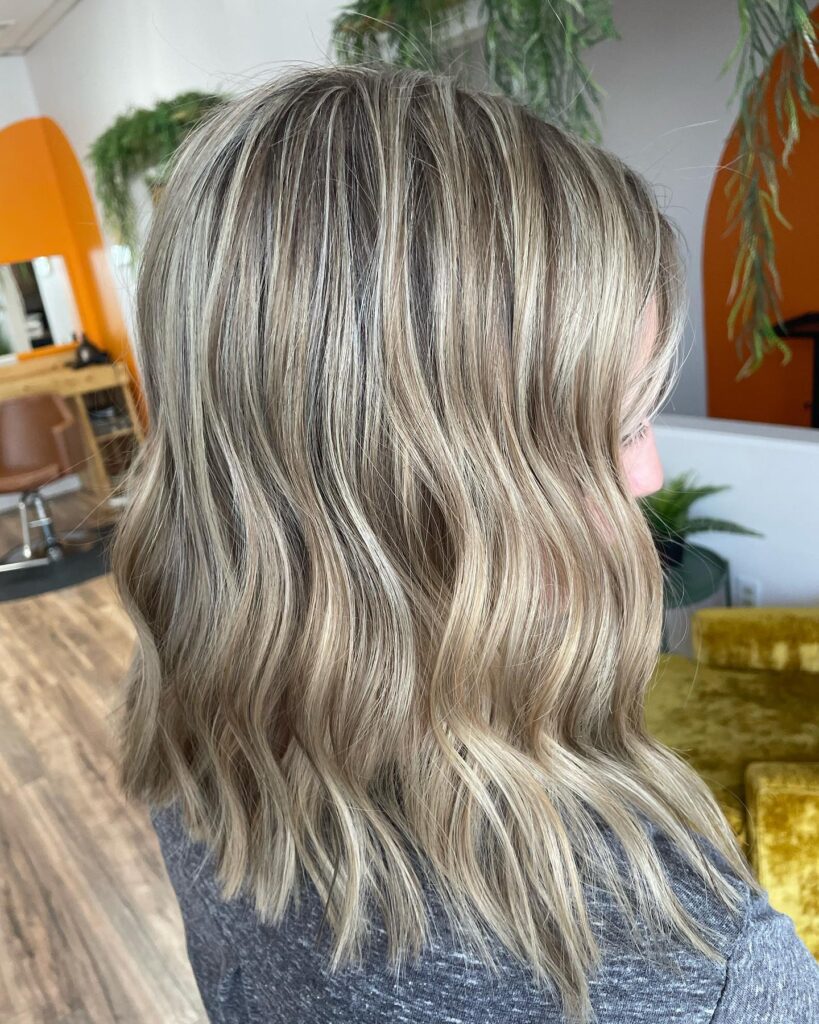 This chic collarbone length lob is styled into the most perfect relaxed beachy waves. The stunning look mixes bright sandy blonde highlights with a cool mushroom blonde base to give it an ultra dimensional finish. It features texturized, blunt cut ends with just a hint of interior layering to keep the movement light, airy, and totally effortless.If you are rocking a rounder face shape, these beautiful vertical wave formations are your absolute best friend. They break up the width of the cheeks and draw the eyes down to instantly give the illusion of a more elongated profile. I love how this easygoing style delivers maximum summer glamour with minimal styling effort.ALT TEXT:
A woman from a side profile view showcasing a shoulder length blonde lob styled in textured beach waves.