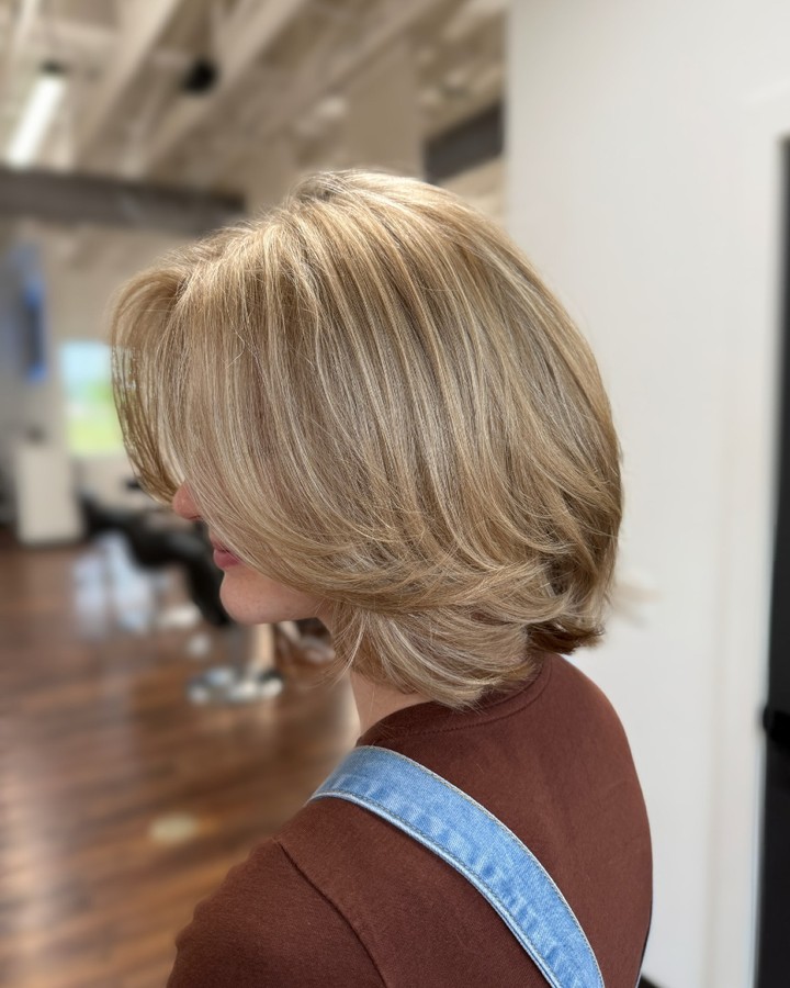A woman seen from a side profile showing a neck length honey blonde bob haircut with soft feathered layers and a blowout style.
