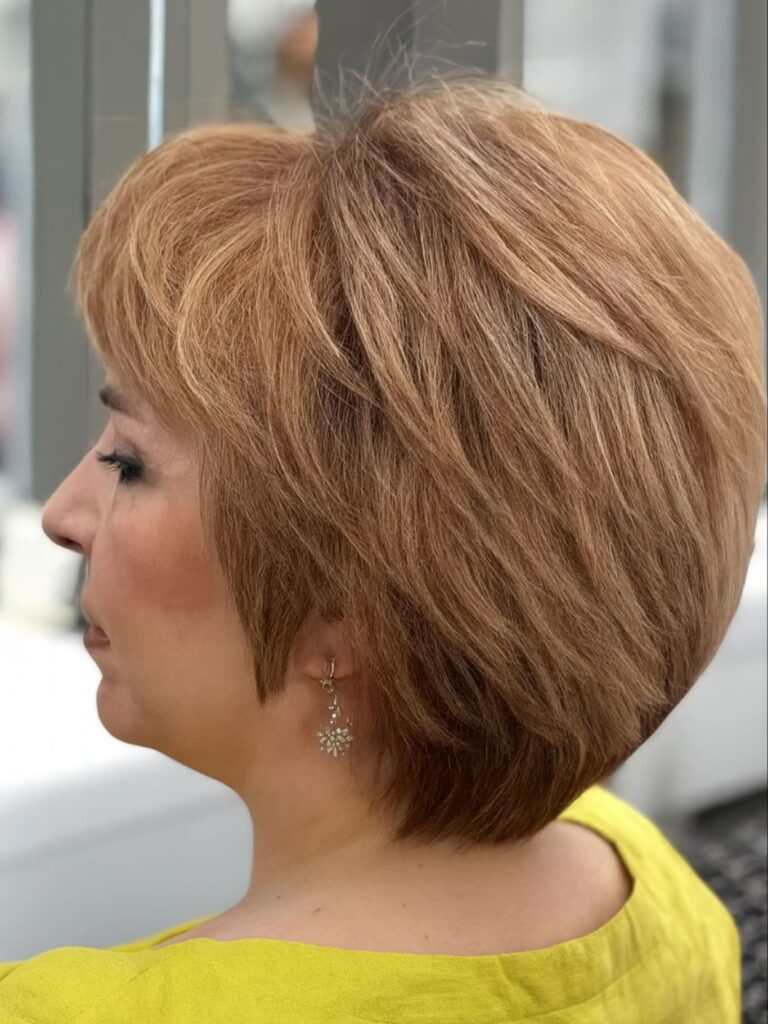A woman with fine hair shows off a voluminous stacked bob in a warm caramel blonde shade with many textured layers.