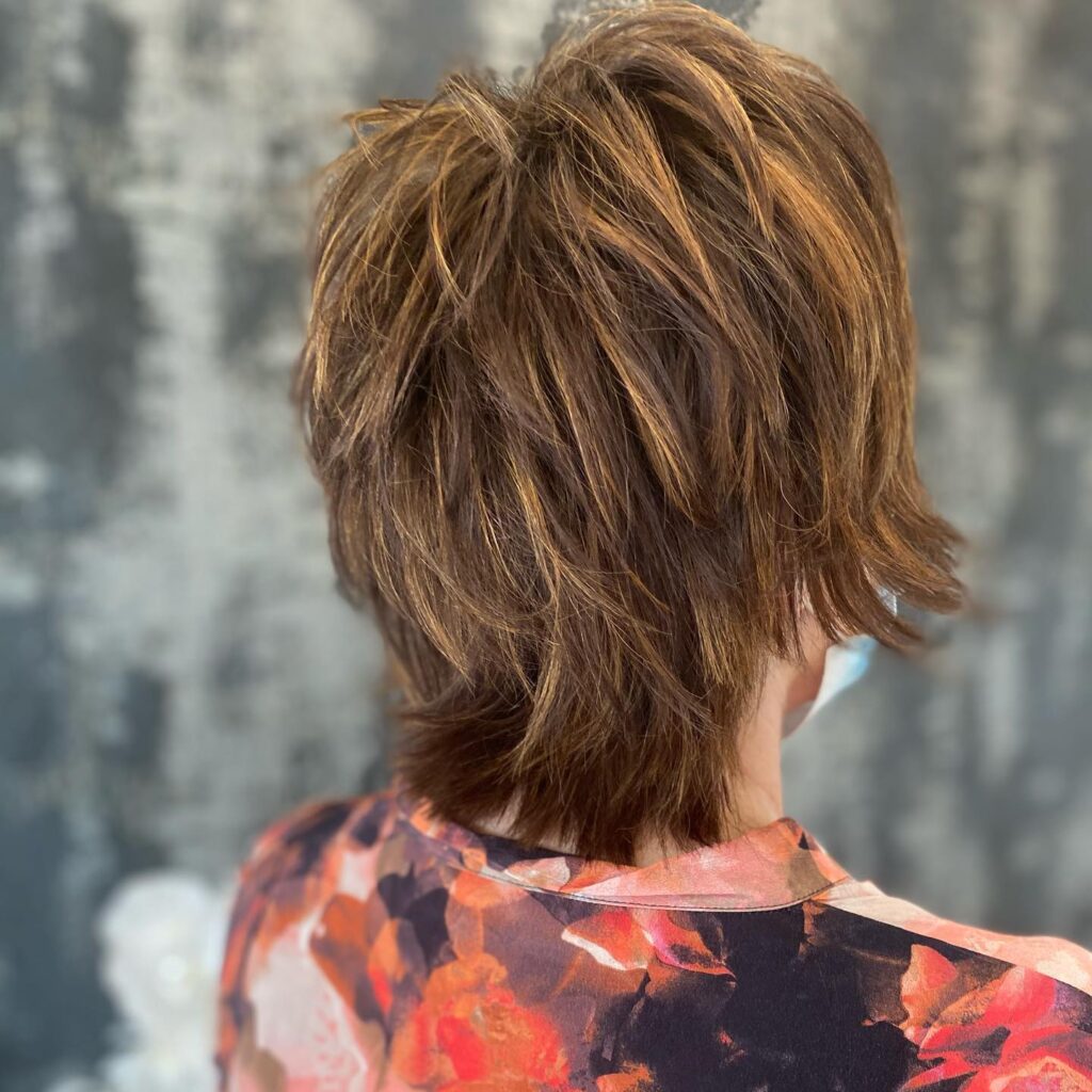 A back view of a woman with a choppy chestnut brown shag haircut featuring golden highlights and many textured layers.