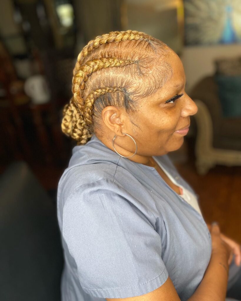 A woman in profile showing thick golden blonde cornrows braided back into a low textured bun.