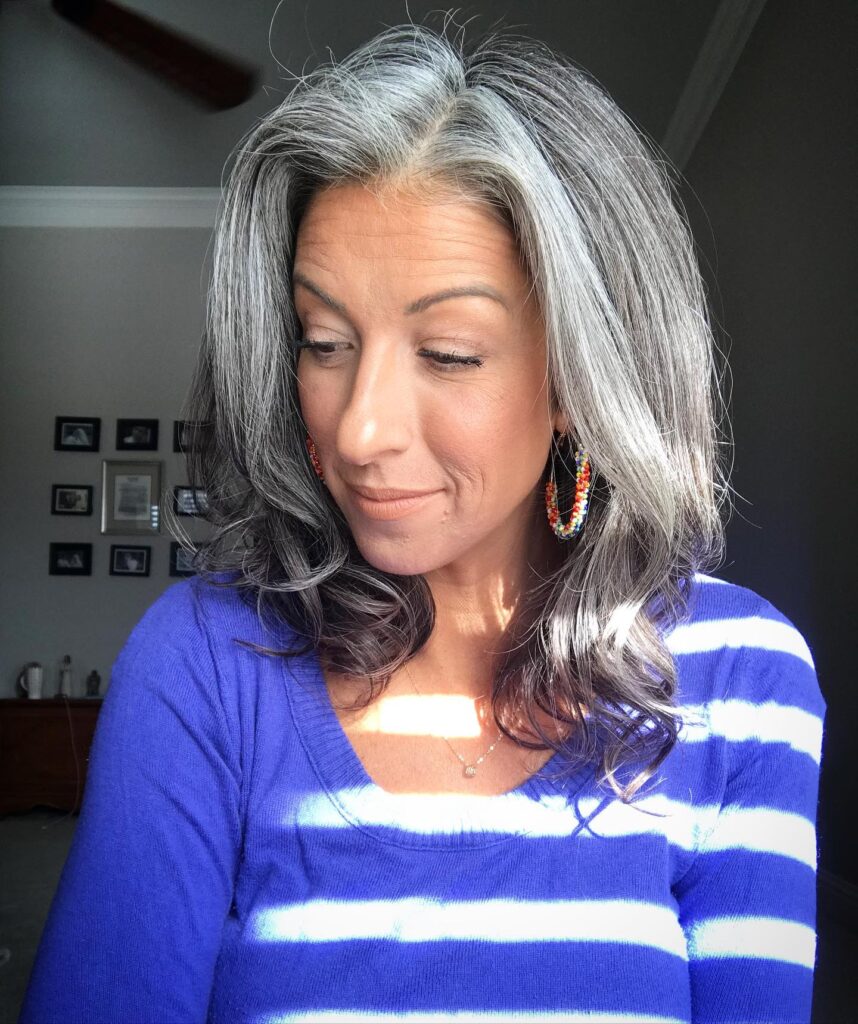 A woman with shoulder length salt and pepper hair styled in soft curls wearing a blue and white striped shirt.