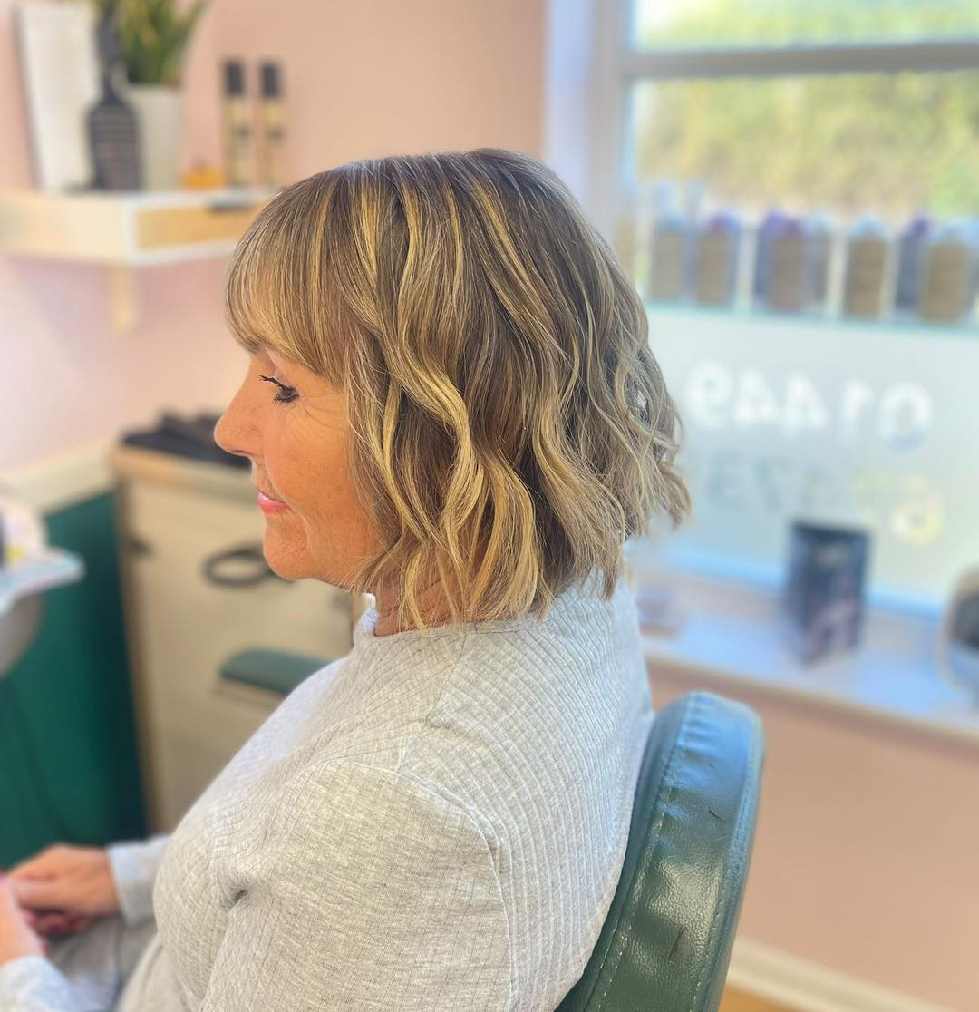 A woman with a blonde wavy bob and full bangs is smiling in a profile view within a salon.