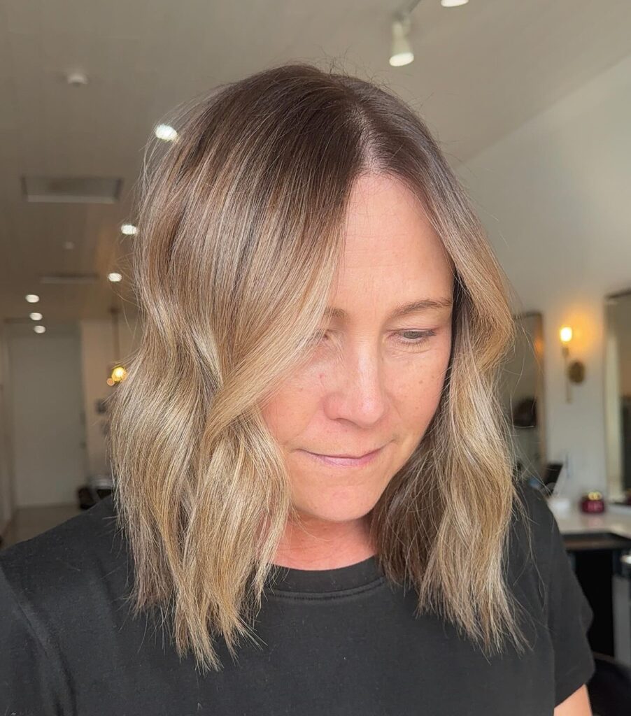 A woman with a middle part showcases a shoulder length sandy blonde lob with soft waves and a natural root shadow.