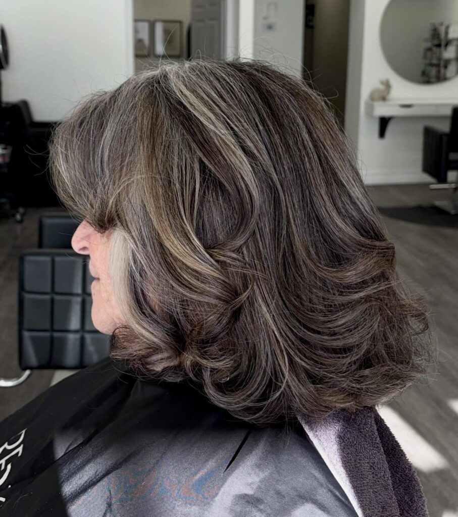 A woman shows off a voluminous shoulder length lob with dark brown hair and bold silver highlights styled in large bouncy curls.