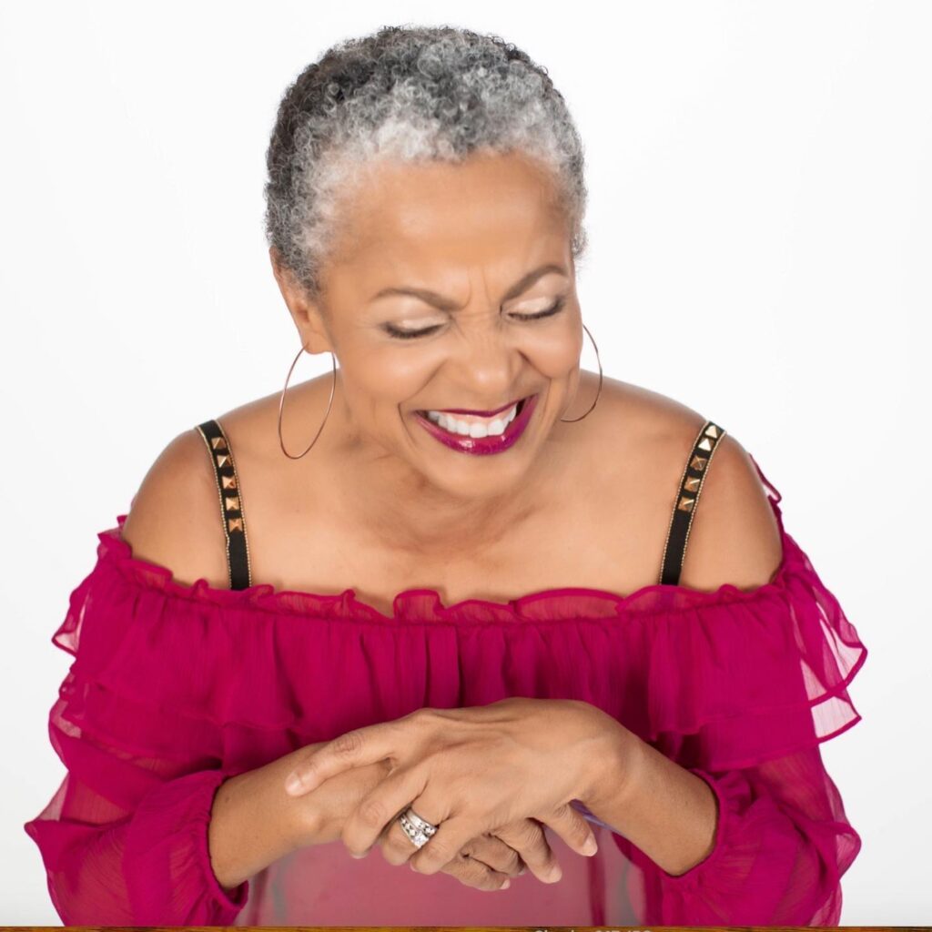 A woman with a joyful expression wearing a very short tapered salt and pepper pixie cut with natural coils.