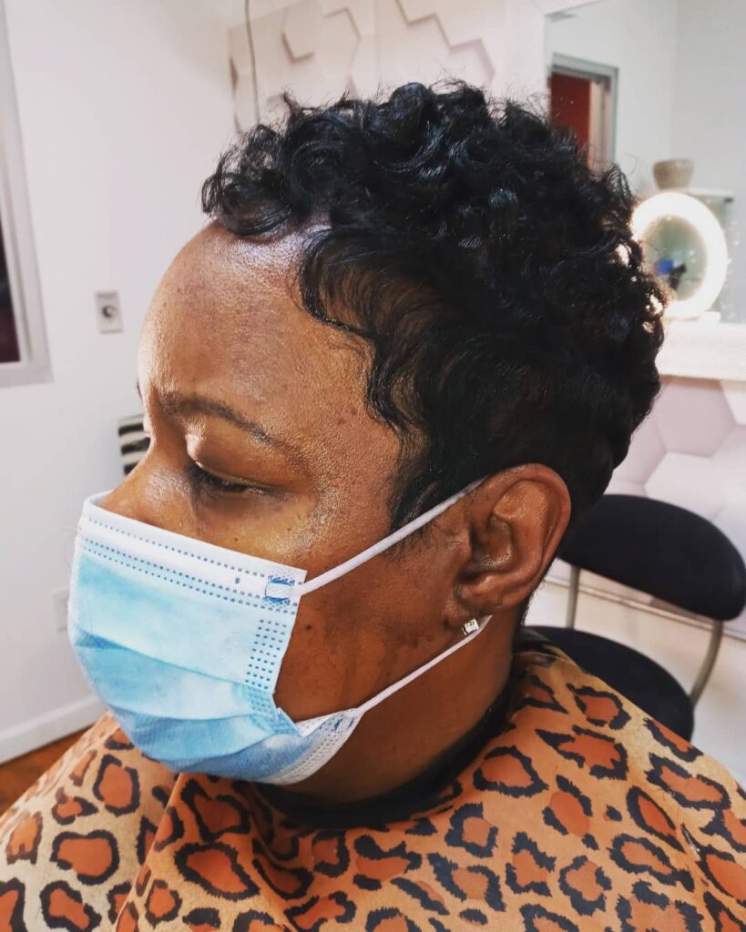A woman with a short textured black pixie haircut featuring tight curls on top and sleek finger waves at the temple.