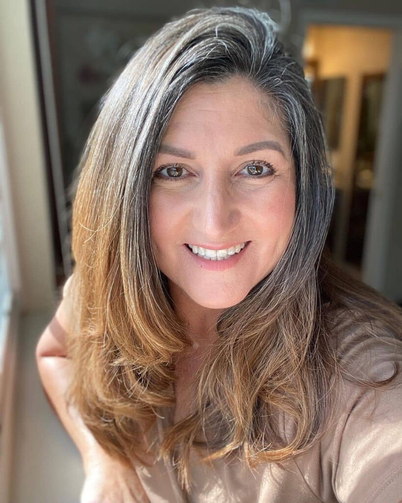 A woman with long layered hair featuring caramel blonde lengths and natural silver roots styled with a side part.