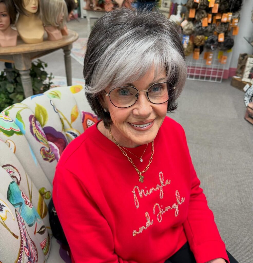 A smiling woman with a long face shape wearing an ear length charcoal and silver layered bob with full bangs and flicked ends.