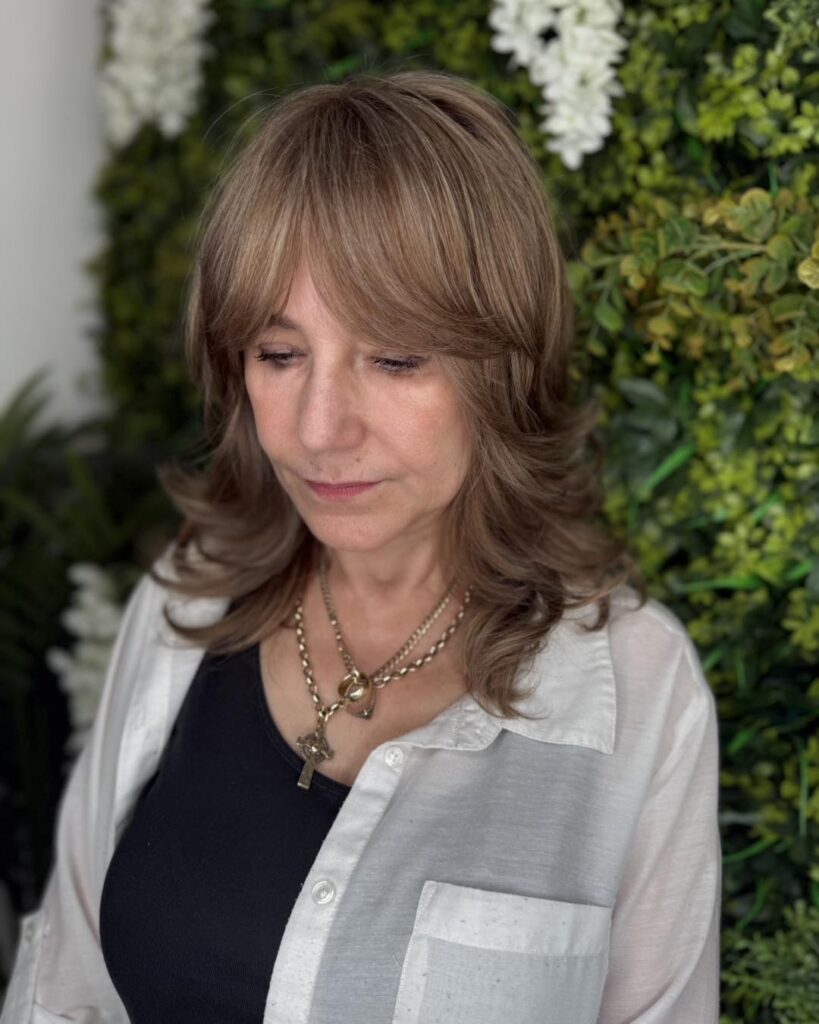 A woman with a long face shape wears a medium length feathery layered brown hairstyle with a full fringe and subtle highlights.