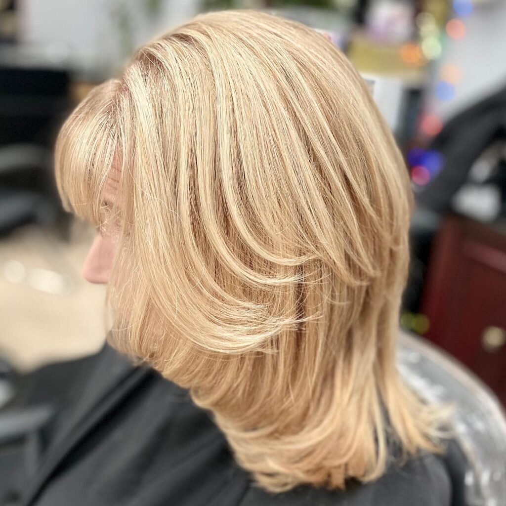 A woman with thick honey blonde hair styled in a mid length butterfly cut with wispy bangs and voluminous layers.
