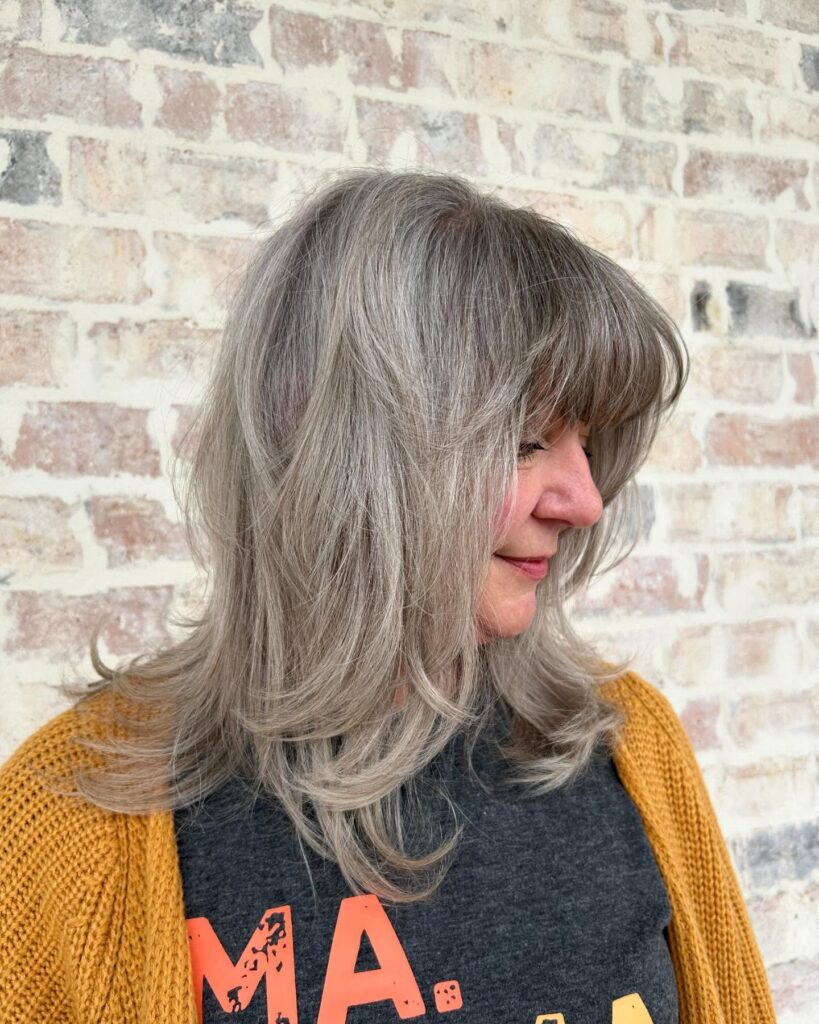 A woman with thick shoulder length silver hair featuring a shaggy layered cut and full bangs.
