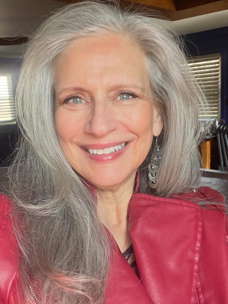 A close up portrait of a smiling older woman with long flowing silver hair layered around her face wearing a red jacket.