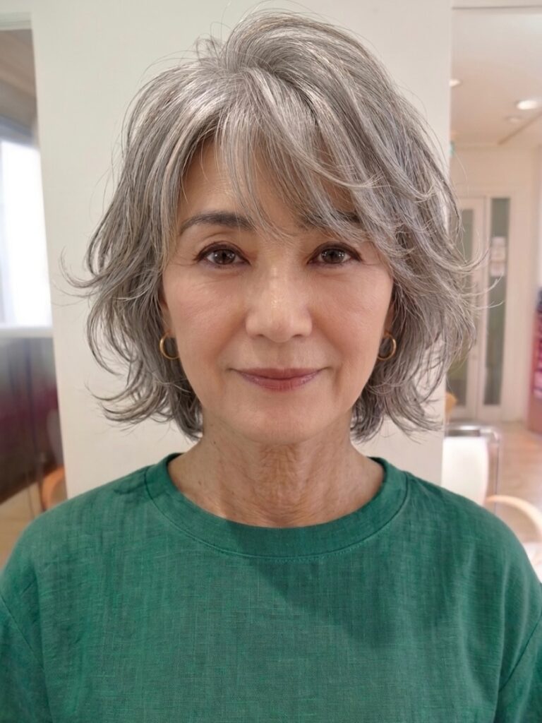 A front facing photo of an older woman with a chin length silver grey layered bob featuring flicked ends and side swept bangs.