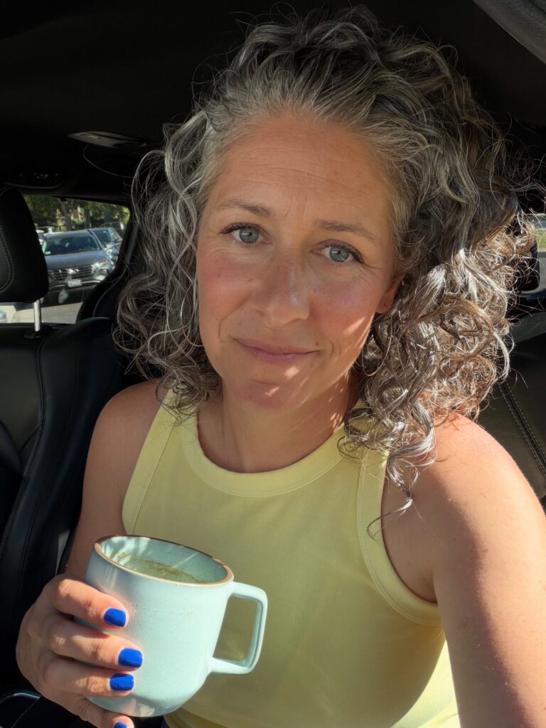 A portrait of a woman with naturally curly salt and pepper hair smiling while holding a coffee cup in her car.