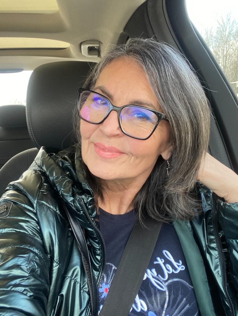 A portrait of a smiling woman with glasses sitting in a car showcasing a collarbone length charcoal lob with subtle silver highlights.