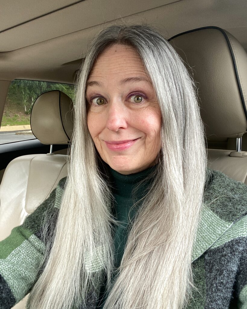 A smiling woman with long straight silver grey hair parted down the middle sitting in a car.