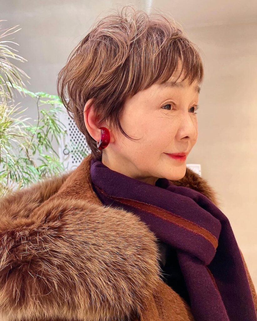 Side profile of an older woman with a chic short textured chestnut pixie cut with wispy bangs and red ear cuffs.