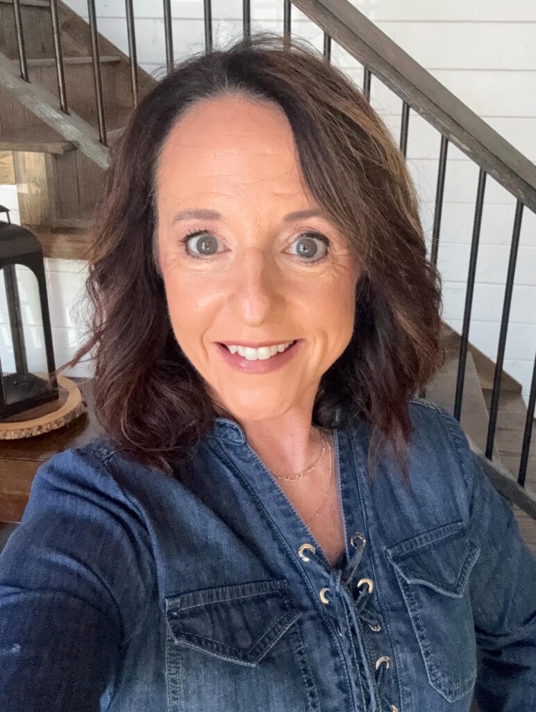 A portrait of a smiling woman with a shoulder length wavy dark brown lob haircut wearing a denim shirt.