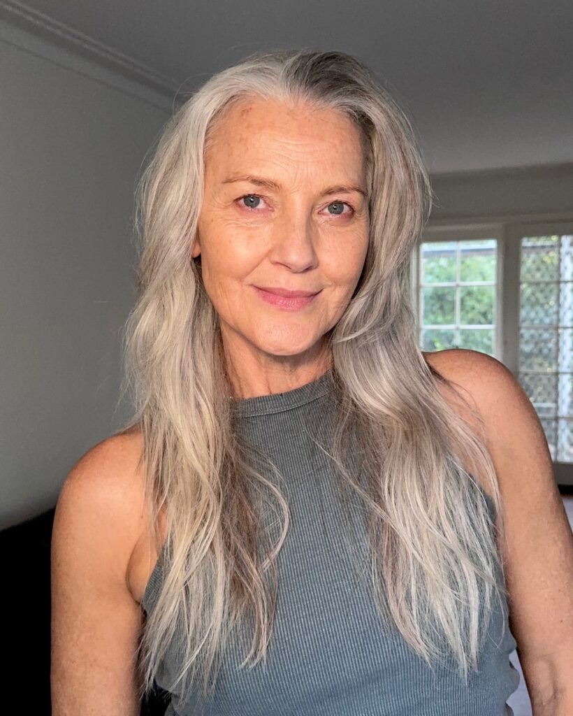 A natural light photo of an older woman with beautiful long silver grey hair with soft waves and face framing layers smiling and looking confidently at the camera.