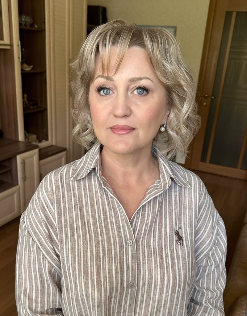 A portrait of an older woman with bright blue eyes and a neck length wavy blonde bob haircut with wispy bangs.