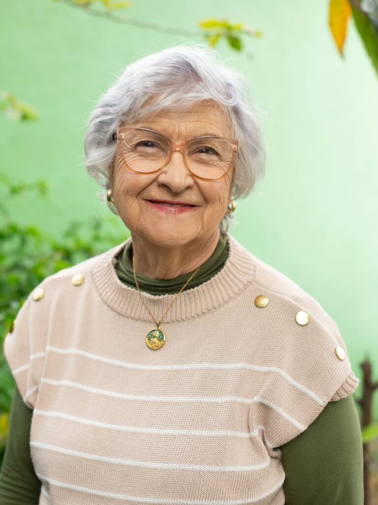 A portrait of a smiling older woman with glasses wearing a beige sweater and a soft feathered silver pixie haircut.