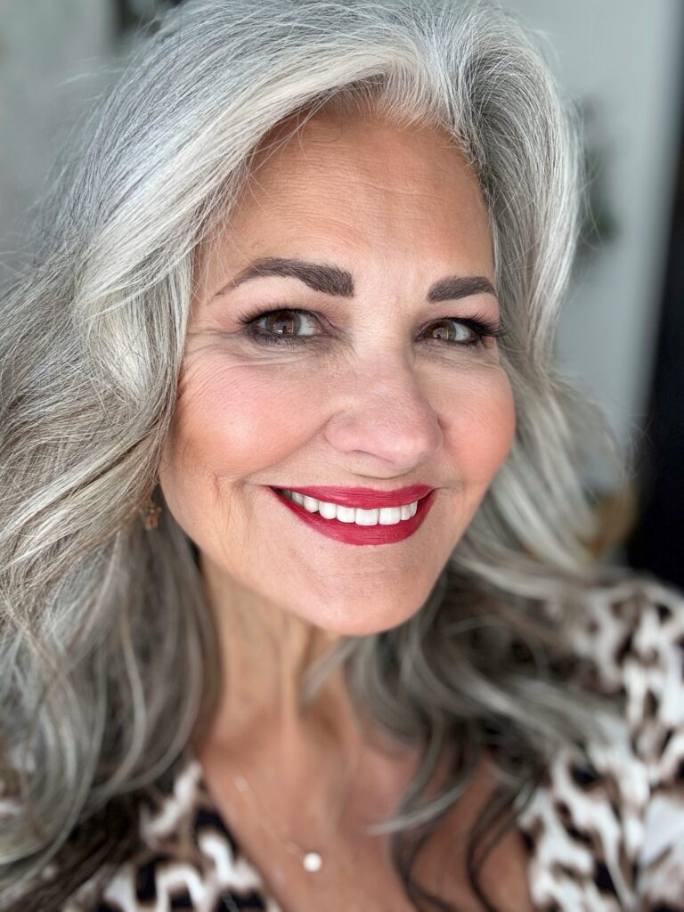 A smiling woman with long layered platinum silver hair and bright red lipstick wearing large sparkly hoop earrings.