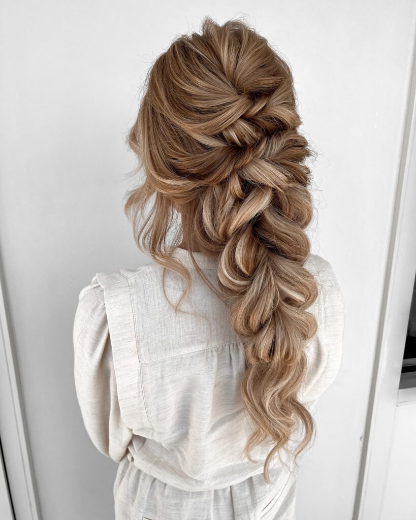 A woman with long highlighted honey blonde hair styled into a thick voluminous boho braid with soft face framing curls.