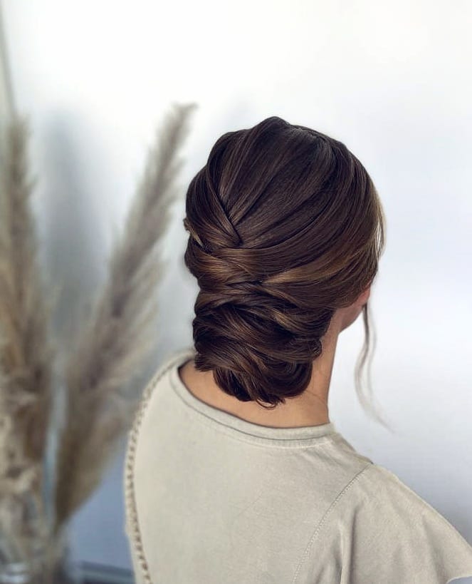 A woman with long dark brown hair styled in a sleek low woven chignon with soft wispy tendrils.