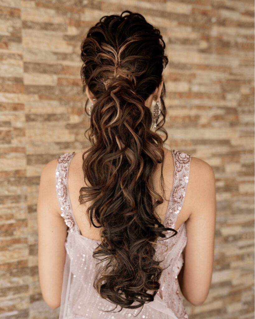 Back view of a woman with long dark brown and highlighted curly hair styled in a textured braided low ponytail.