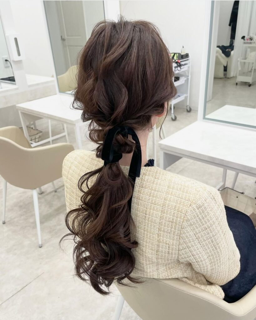 A woman with long dark brown wavy hair styled in a low ponytail wrapped with a black velvet ribbon.