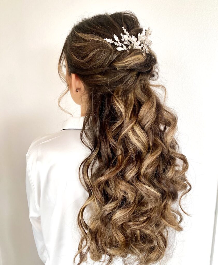 A woman with long highlighted brown hair styled in a romantic half up twisted look with voluminous waves and a crystal floral hair accessory.